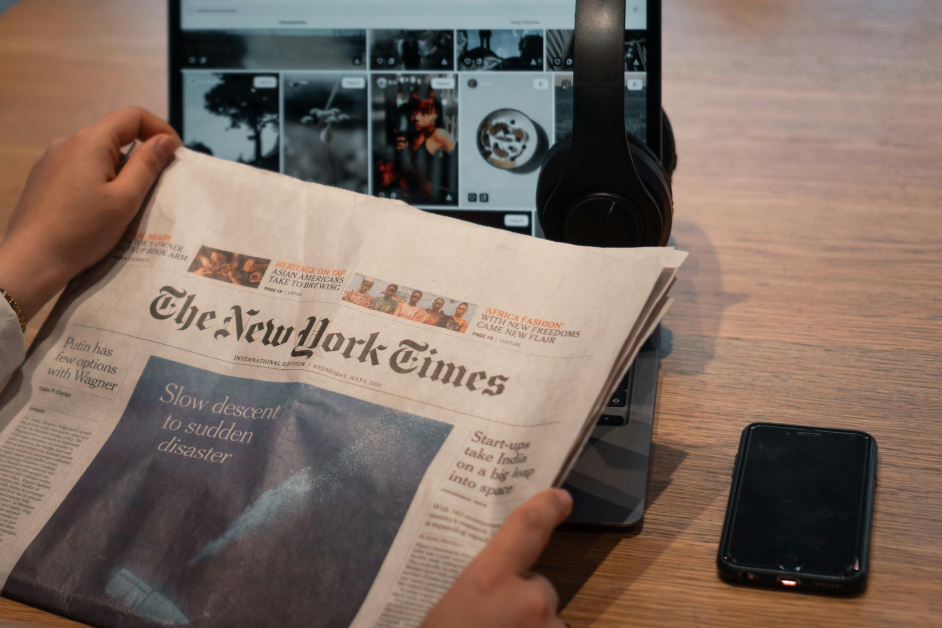 Close-up of hands holding a newspaper by a laptop and smartphone on a table.