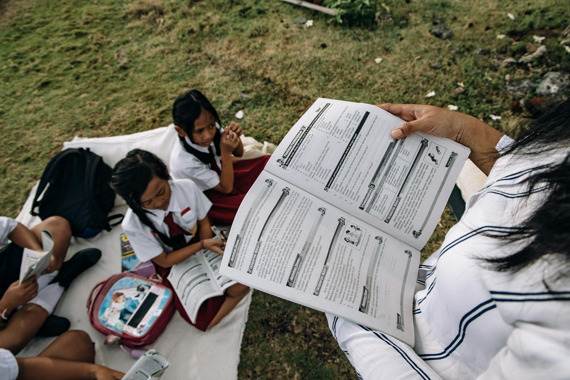Group of school children in uniforms reading outdoors on a grassy area.